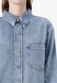 Denim shirt in light blue with a button-up front, pointed collar, left chest pocket, and visible stitching details.
