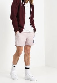 Lyhythihainen valkoinen t-paita marsalanpunaisen hupparin alla, yhdistelmä vaaleanpunaiset shortsit, joissa on tummansininen logo. Valkoiset lenkkarit mustasuorilla sukilla.