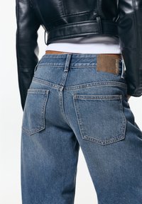 Modré deninové džínsy so strihom rovný nohavic, s dvoma zadnými vreckami a hnedou koženou nášivkou na opasku. Klasická vyšívacia detail.