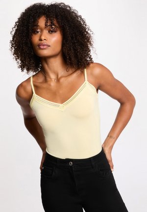 Donna con capelli ricci indossa una canottiera giallo pallido con rifiniture in pizzo e pantaloni neri, in piedi con le mani dietro la schiena davanti a uno sfondo bianco.