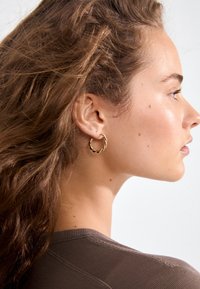 Gold hoop earring featuring a twisted design; small and lightweight, worn on the left ear, with a smooth finish and bright metallic sheen.