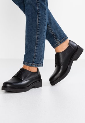 Personne portant un jean bleu et des chaussures en cuir noir à lacets, debout sur un sol blanc avec un pied légèrement levé.