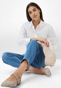 Beige Wildlederballerinas mit gerafften Akzenten, getragen mit blauen Jeans mit umgeschlagenem Saum und einem weißen Hemd mit Knöpfen. Eine beige Kappe liegt in der Nähe.
