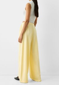 Yellow wide-leg trousers made of smooth fabric, featuring pleats at the front and a high waist, paired with a ribbed white top.