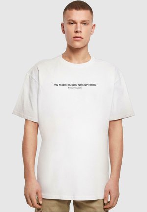 White cotton t-shirt with oversized fit, featuring black text: "YOU NEVER FAIL UNTIL YOU STOP TRYING #NeverGiveUp" across the chest.
