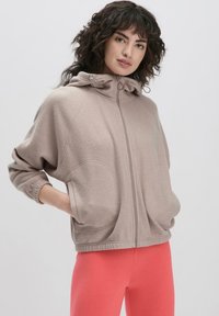 Lichtbeige ritsvest met een gestructureerde, zachte stof, een capuchon met trekkoord en grote zijzakken. Gecombineerd met felroze legging.