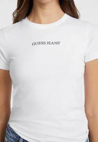 Witte katoenen t-shirt met korte mouwen en een ronde hals. Heeft het "GUESS JEANS" logo in het zwart op de borst. Glad textuur.