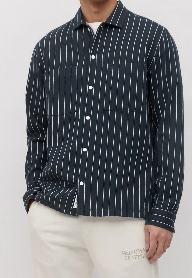 Homme portant une chemise bleu marine à manches longues avec des rayures verticales blanches et une fermeture à boutons, associée à un pantalon crème portant le texte « Marc O'Polo CRAFTED ».