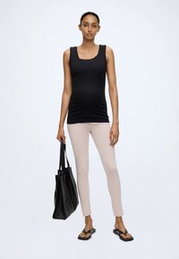 Kvinde står vendt fremad iført sort tanktop, lyse beige leggings, sorte sandaler og holder en stor sort mulepose.