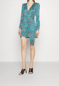 Tee-farbendes Blumenkleid mit langen Ärmeln, tailliertem Bund und asymmetrischem Saum. Schwarze offene, hochhackige Mules. Glatte Stofftextur.