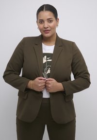 Hnědý propracovaný blazer přes bílý tričko s potiskem rostliny. Hladká látka s strukturovanými rameny a klopy. Viditelný zlatý náramek.