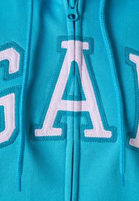 Jasnoniebieska bluza z kapturem na zamek błyskawiczny, z dużymi białymi naszywanymi literami "GAP" na przodzie, o gładkiej fakturze i dopasowanym zamku.