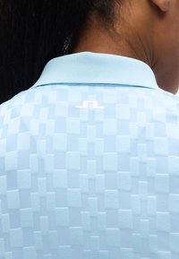 Ανοιχτό μπλε πόλο πουκάμισο με υφή τετραγώνων, γιακά και μικρό λευκό λογότυπο στην πλάτη. Λείο ύφασμα.