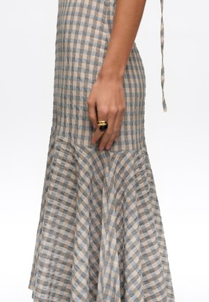 Mano apoyada en el lateral de un vestido de cuadros beige y gris con dobladillo acampanado, llevando un anillo llamativo grande de oro y negro en el dedo medio.