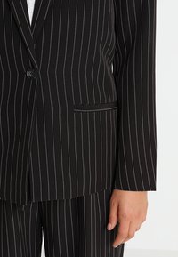 Blazer noir à fines rayures en tissu texturé, avec fermeture à boutons, revers crantés et une poche avant unique inclinée.