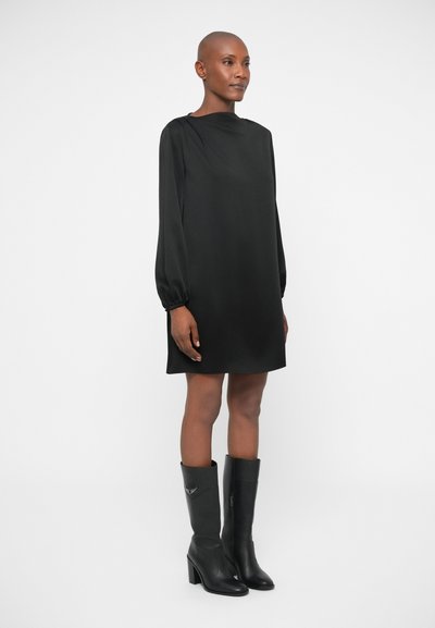 Rochie neagră cu mâneci lungi, guler înalt și croială lejeră, asortată cu cizme din piele negre, cu toc, până la genunchi, care au un design subtil.