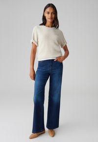 Beige, strukturierter Strickpullover mit aufgerollten Ärmeln, kombiniert mit weit geschnittenen, dunkelblauen Jeans. Spitz zulaufende, beige Ballerinas runden das Outfit ab.