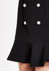 Svart klänning med en utsvängd nederkant, strukturstoff och prydd med sex cirkulära pärlliknande detaljer längs framsidan.