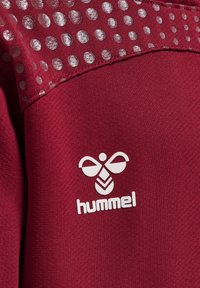 Burgundy idrottstopp med en slät textur, med en silverprickig design på axeln och en vit "hummel"-logotyp på framsidan.