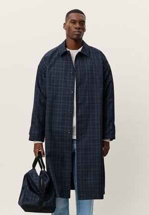 Navy plaid long coat made of a smooth fabric, featuring a button closure, wide collar, and side pockets. The model holds a black duffel bag.
