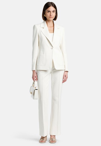 Witte op maat gemaakte blazer met grote opgestikte zakken, gecombineerd met bijpassende broeken. Model houdt een kleine witte handtas vast. Lichte, strappy hakken.