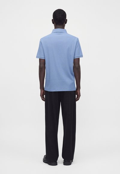 Homme debout de dos, portant un polo bleu clair à manches courtes, un pantalon noir à jambes larges et des chaussures noires sur un fond uni.
