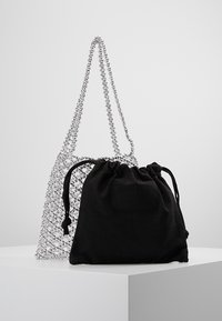 Sac à main transparent en perles avec des anses en chaîne argentées et une pochette noire à cordon tirée reposant contre lui sur un piédestal blanc.