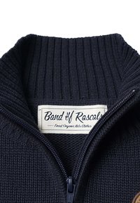 Sini sininen neulepaita, jossa on korkea kaulus ja täysi vetoketju. Ominaisuuksina ribbikuviointi ja etiketti, jossa lukee "Band of Rascals."