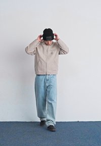 Giacca beige con tasca frontale e logo discreto, abbinata a jeans larghi di un leggero blu e un berretto nero. Scarpe grigie completano il look.
