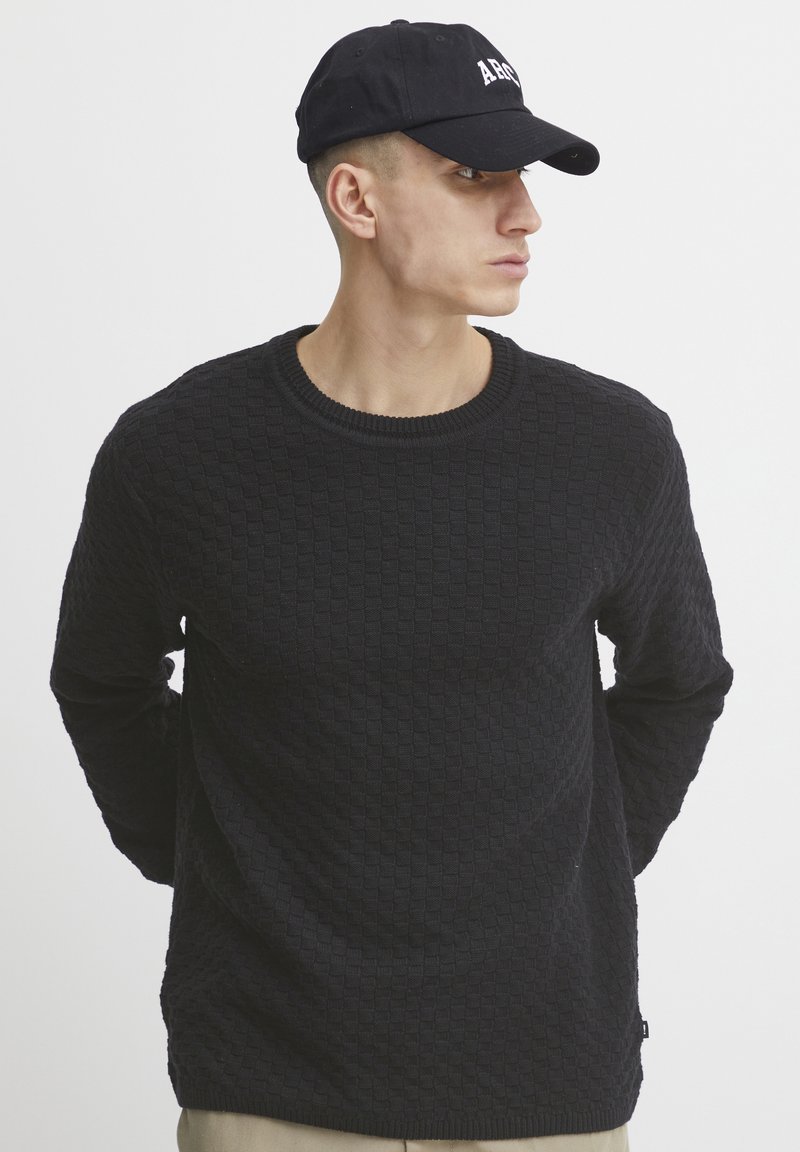 Black textured knit sweater with a round neck, worn by a person, paired with a black cap featuring white lettering.