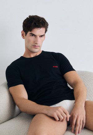 Mann mit dunklen Haaren, der ein schwarzes Hugo-T-Shirt und weiße Shorts trägt, sitzt auf einem beigen strukturierten Sessel vor einer schlichten weißen Wand.