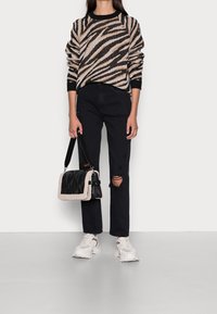 Femme portant un pull brun à rayures zébrées noires, jean noir déchiré, baskets blanches, tenant un sac noir avec garniture en fourrure beige.