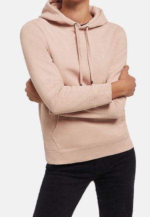 Personne portant un sweat à capuche rose clair avec cordons, bras croisés, associé à un pantalon noir, sur un fond uni.