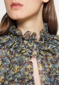 Nahaufnahme vom Hals und den Lippen einer Frau, die eine mehrfarbige, gerüschte Bluse mit blauen, gelben und braunen abstrakten Blumenmustern trägt.