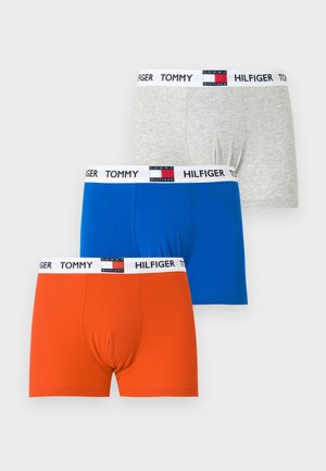 Jeune homme debout pieds nus portant un boxer bleu Tommy Hilfiger sur un fond blanc uni.