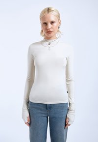 Light beige long-sleeve fitted top with a high neck, made of smooth material, featuring thumb holes and a plain design. Paired with blue jeans.