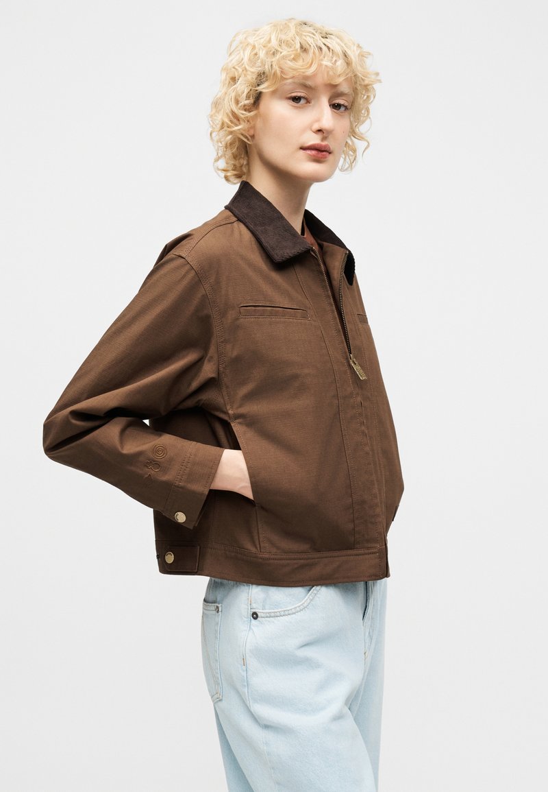 Persona con capelli ricci corti e biondi indossa una giacca marrone con colletto nero e jeans azzurri, in piedi con una mano in tasca.