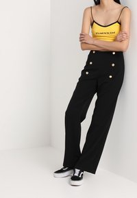 Personne portant un crop top jaune avec le texte « C'est cool d'être gentil », un pantalon noir taille haute avec des boutons dorés et des baskets noires.