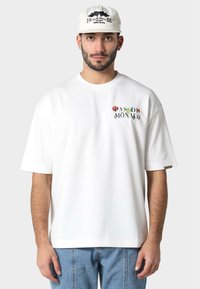 Balta medvilnės T-shirt marškiniai trumpomis rankovėmis, su spalvingu gėlių užrašu "PAS DE MONACO" priekyje; laisvas kirpimas. Dėvimi su šviesiai mėlynomis džinsomis.