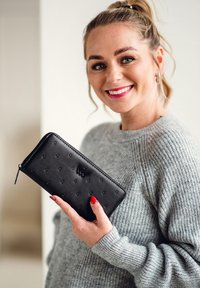 Black leather wallet with embossed dots, zip closure, and logo detail. Held in a hand wearing a gray sweater, displaying red nail polish.