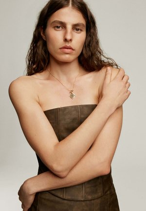 Joven mujer con cabello largo y rizado, vistiendo un top marrón sin tirantes y un collar con colgante dorado, sujetándose el hombro opuesto.