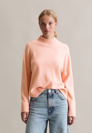 Perzikkleurige gebreide trui met een hoge hals, drop shoulders en geribde manchetten. Gecombineerd met lichtblauwe high-waisted jeans. Zachte textuur.