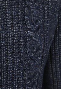 Close-up van donkerblauwe gebreide stof met ribbel- en gevlochten kabelpatronen en witte stippen die erin geweven zijn.