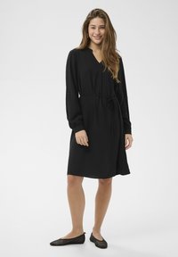 Rochie neagră cu mâneci lungi, decolteu în formă de V și talie cu cordon. Material texturat, lungime până la genunchi, asortată cu pantofi comozi negri din plasă. Design simplu și versatil.