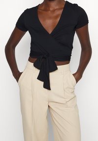 Černý zavinovací top z měkké látky s krátkými rukávy, hlubokým výstřihem do V a zavazovacím detailem v pase, v kombinaci s krémovými kalhotami.
