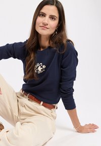 Granatowy sweatshirt z długim rękawem z haftem w kwiaty, połączony z beżowymi spodniami z wysokim stanem i brązowym paskiem. Tekstura jest gładka.