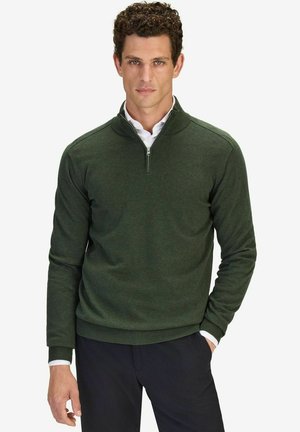 Groene zip-neck trui van gestructureerde brei stof, met een geribbelde zoom en manchetten. Gecombineerd met een witte blouse, tegen een effen achtergrond.