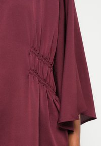Blusa bordeaux con maniche rilassate e oversize e texture arricciata lungo i lati. Il tessuto appare morbido e leggero.
