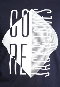 Černé bavlněné tričko s velkým bílým geometrickým designem, na levé straně s textem "CORE" v tučném písmu a na pravé straně s textem "JONES" v lineárním vzoru.