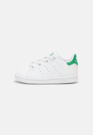 Biała tenisówka Adidas Stan Smith z zielonym elementem na pięcie, perforowanymi bokami i sznurowanym zapięciem, przedstawiona z profilu na białym tle.